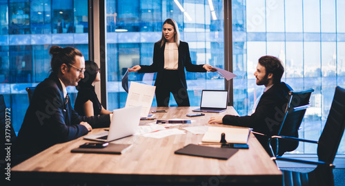 Puzzled female director in elegant suit communicating with employees during working process in conference room standing near meeting table with modern technology, laptop with mock up for website