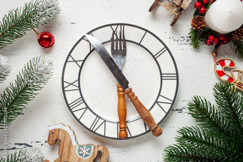 White plate with the clock and cutlery in a frame of their branches of a fir on a white wooden background. Christmas concept. Top view. Flat lay