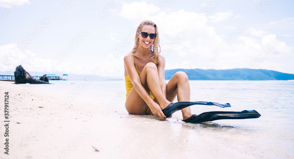 Young blonde in sunglasses putting on flippers on beach