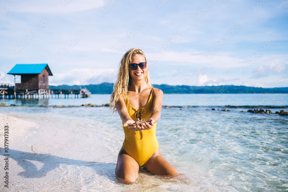 Portrait of funny tan female in trendy sunglasses smiling at camera satisfied with collecting hobby and summer paradise travelling, happy woman 20s with shell mollusk posing at coastline beach