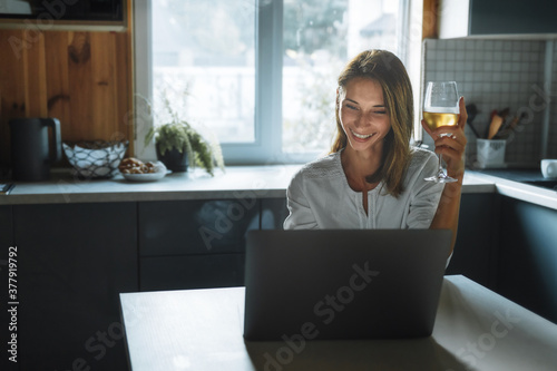 Dating online concept. Happy young woman with glass wine talking through social network sitting at home in the kitchen. Search love, partner and girlfriend on the Internet