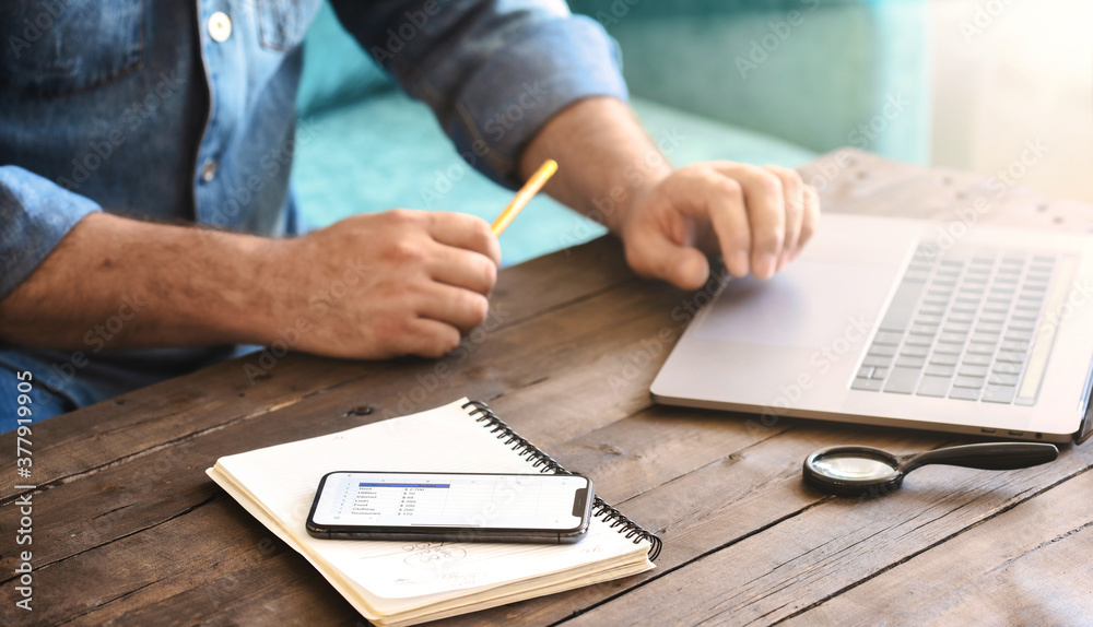 Man sitting on the sofa at home examining home finance. Smartphone with home monthly budget list on wooden table Man sitting on the sofa at home examining home finance. Smartphone with home monthly budget list on wooden table