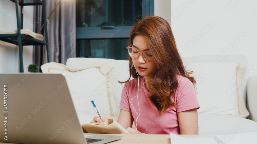 Young Asia teen girl student wears headphones distance learning lesson with online teacher on laptop in living room from home at night. Social distancing, quarantine for corona virus prevention. Young Asia teen girl student wears headphones distance learning lesson with online teacher on laptop in living room from home at night. Social distancing, quarantine for corona virus prevention.