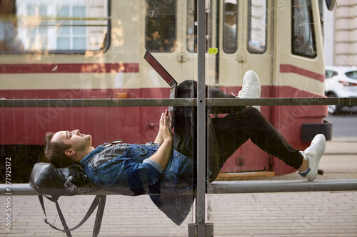 Man lies on bus stop with laptop.