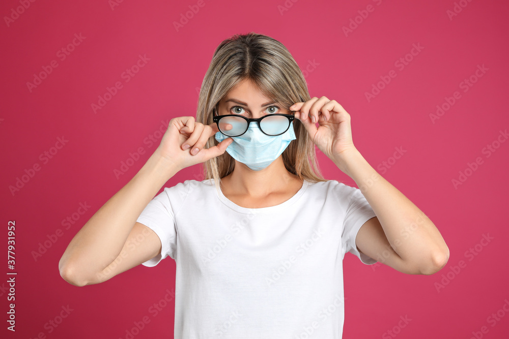 Woman wiping foggy glasses caused by wearing disposable mask on pink background. Protective measure during coronavirus pandemic Woman wiping foggy glasses caused by wearing disposable mask on pink background. Protective measure during coronavirus pandemic