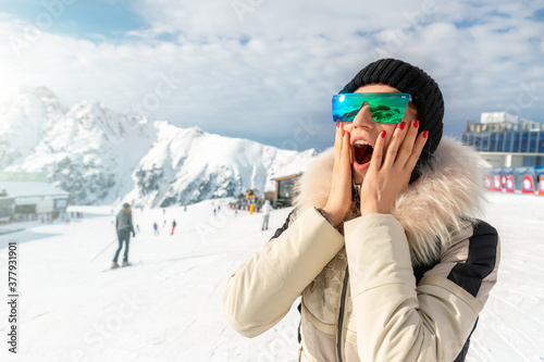 Portrait of beautiful young adult caucasian woman in sport suit, hat, sunglasses smiling looking aside and making excited amazed face expression on mountain peak sunny winter day alpine ski resort