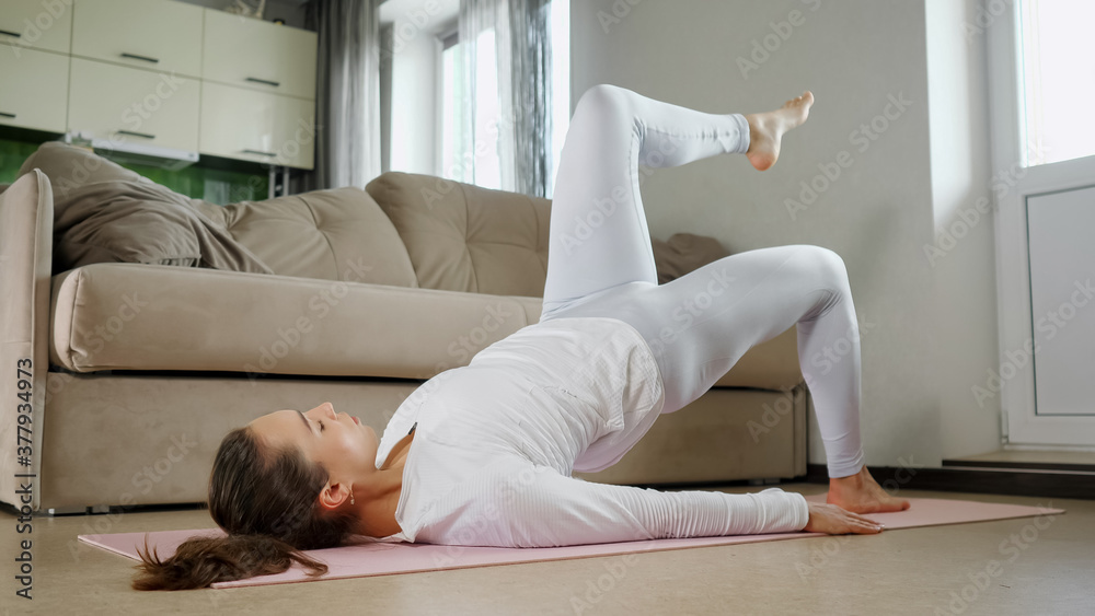 young brunette woman in stylish white tracksuit does sports exercises lying on pink mat and lifting leg near large couch in light room at home
