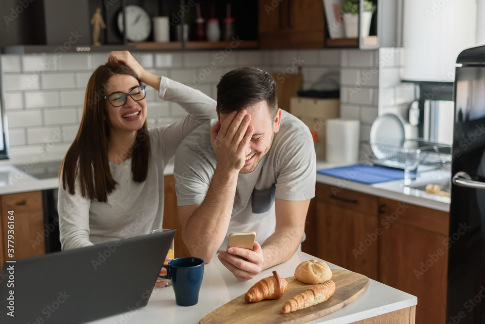 Smiling couple enjoying a beautiful morning at home kitchen, having breakfast, drinking coffee and juice, and looking at smartphone Smiling couple enjoying a beautiful morning at home kitchen, having breakfast, drinking coffee and juice, and looking at smartphone