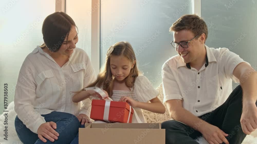 Adorable Girl Unpacking the Order. Happy Family Unpacking the Order. A Young Girl Takes From the Carton Box a Red Present Adorable Girl Unpacking the Order. Happy Family Unpacking the Order. A Young Girl Takes From the Carton Box a Red Present
