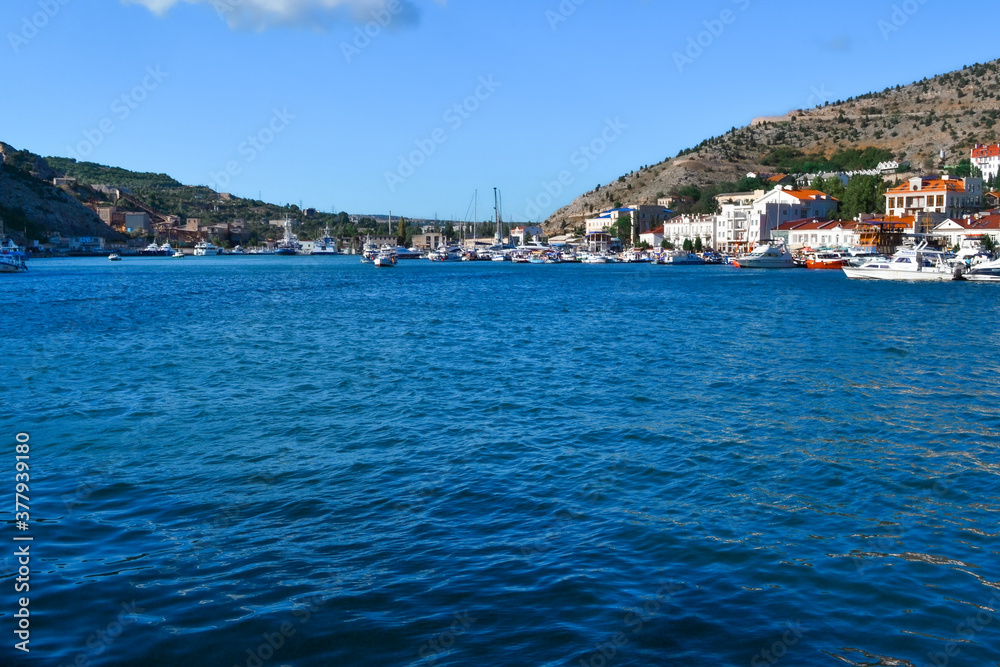 blue water with waves in bay of mountain town of Balaklava with marinas, ships, boats and bright buildings, ripples, sunny blue water with waves in bay of mountain town of Balaklava with marinas, ships, boats and bright buildings, ripples, sunny