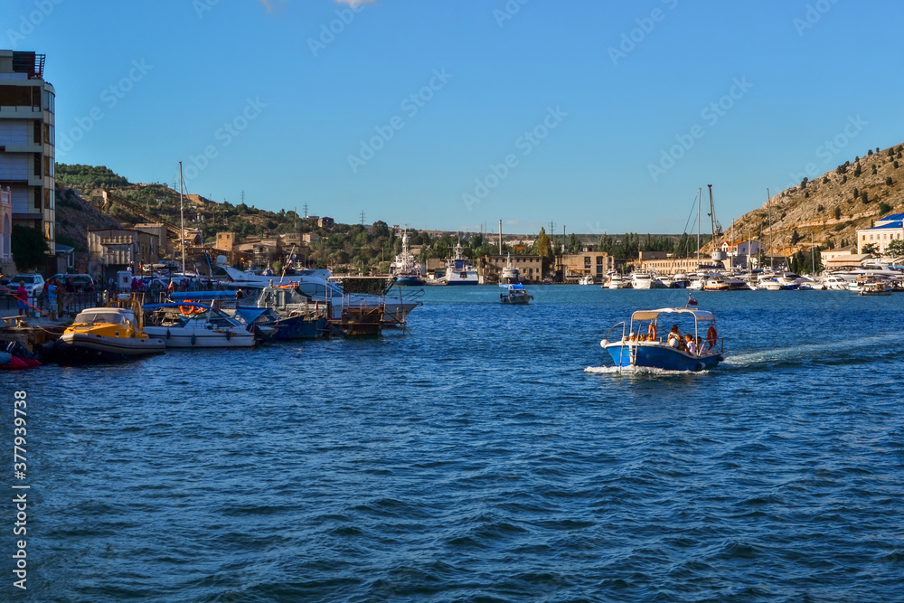 bright blue boat along the bay of resort town of Balaklava among white ships on docks near houses and buildings, sunny