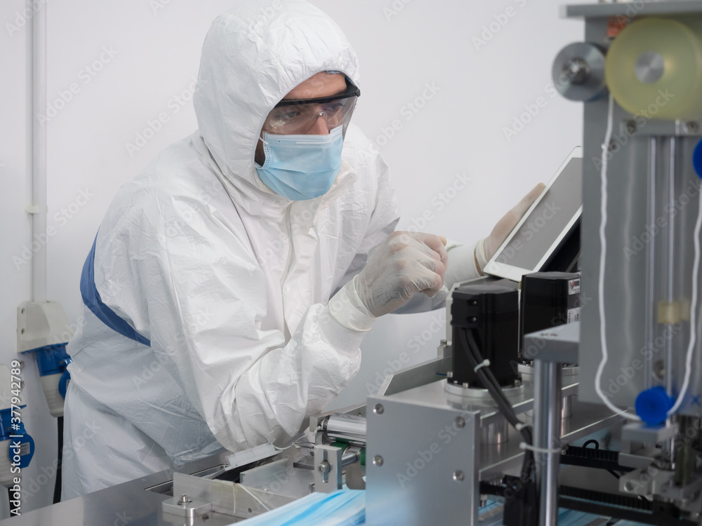 Naklejka premium The engineer worker in protective suits, masks, and gloves is checking and controlling the machine in the manufacture of surgical masks for coronavirus..