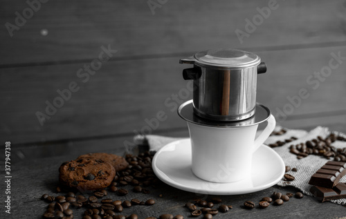 White cup of coffee with a metal filter on a top and scattered beans on a wooden table. Traditional vietnamese coffee.