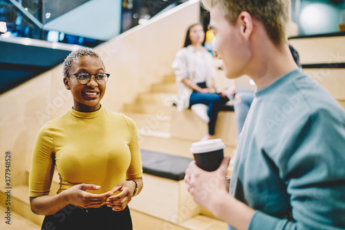 Smiling dark skinned woman in eyewear talking with japanese male colleague discussing startup project together, positive male and female employees having friendly communication on coffee break