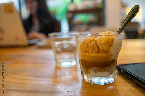 Cold Brew Iced Coffee Affogato, Vanilla Ice-Cream in a glass next to smart phone on wooden table and customers using laptop for work in blurred background.