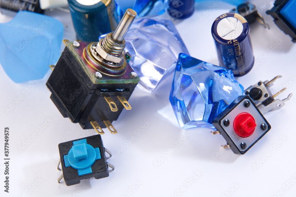 Closeups of capacitors and switches set against a white background with blue marbles and crystals in the foreground. Closeups of capacitors and switches set against a white background with blue marbles and crystals in the foreground.