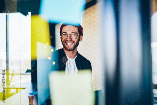 Cheerful caucasian hipster guy in trendy eyewear satisfied with creative job laughing sitting near stickers with ideas in office, 20s smiling carefree male student enjoying learning in business school photo