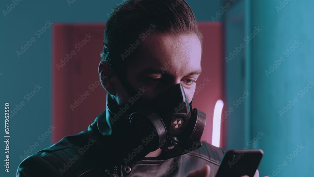 Portrait of brutal man in respirator using smartphone at night over neon lights. Cyberpunk concept. Male in protective mask using mobile app on blue and pink neon light background Portrait of brutal man in respirator using smartphone at night over neon lights. Cyberpunk concept. Male in protective mask using mobile app on blue and pink neon light background