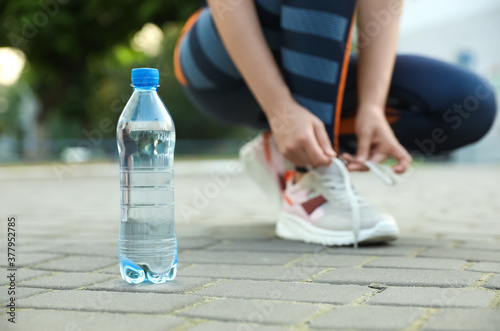 Closeup view of sportswoman lacing sneakers outdoors, focus on bottle with water