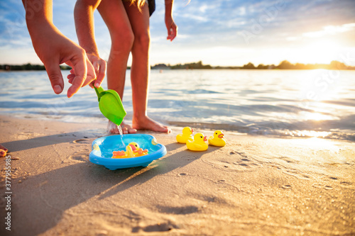 Side crop view of unrecognizable caucasian girl playing with rubber yellow ducks in small blue pool, standing on beach sand. Incognito kid having fun, enjoying holidays at seashore, summer sunset.