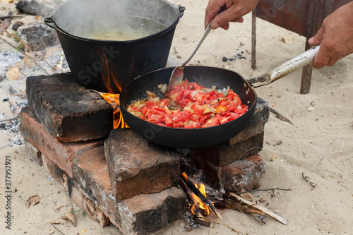 man's hands coock in black iron pan roasting vegetables. big cooking pot with soup stand in camping fire and getting prepared for outdoor dinner. Camping picnic