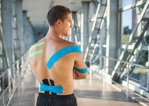 Back view of muscularman wearing black sports outfit  looking aside. Young male athlete posing indoors  colorful kinesiology taping on naked back  futuristic interior. Sports rehabilitation concept.
