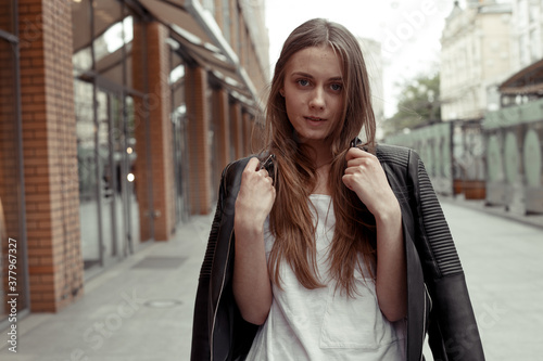 The middle shot portrait girl with leather black jacket draped over her shoulders posing at the camera. Female model standing in the middle of the street and looking into the frame