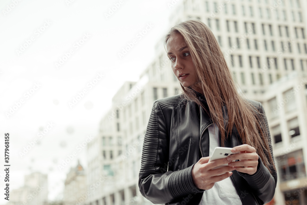 Confident woman wearing black leather jacket and white shirt holding mobile phone on a urban background. Confident woman wearing black leather jacket and white shirt holding mobile phone on a urban background.