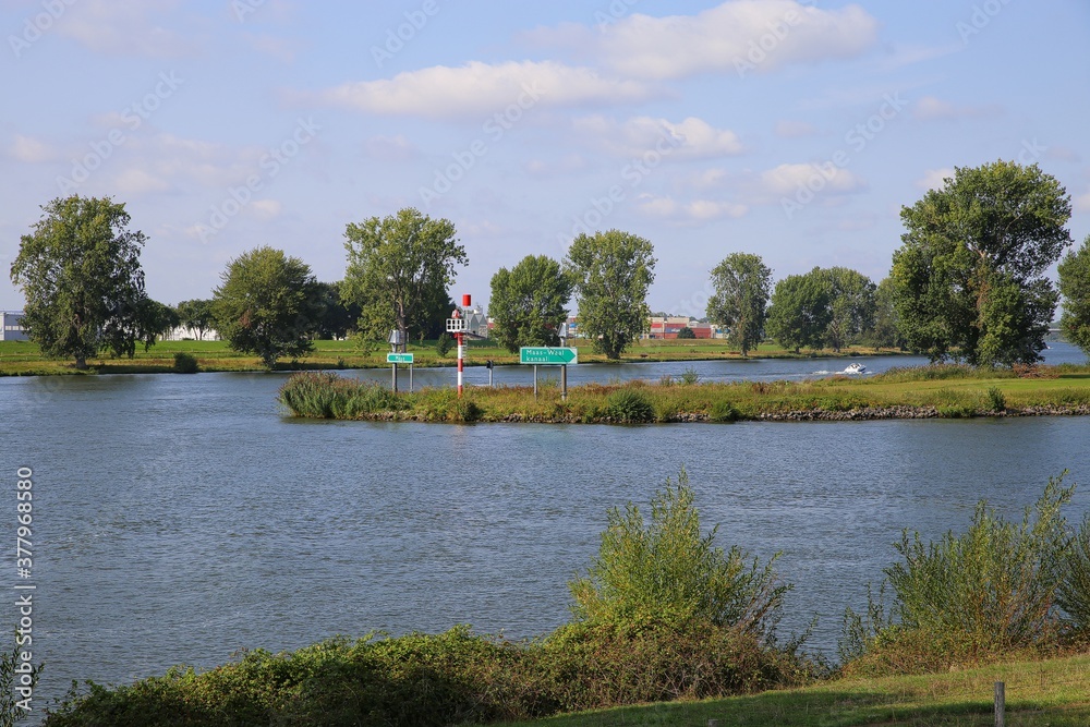 View on junction of river maas with in rural area with rhine maas canal sign (dutch text on sign: maas waal canaal) from Heumen to Nijmegen - Netherlands