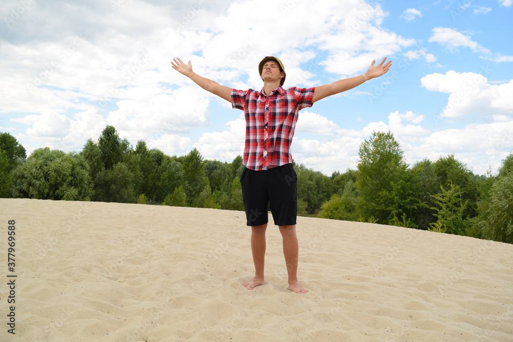 Obraz premium A young man in a bright checkered T-shirt stands with raised hands in nature