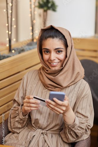 Vertical portrait of young Middle-Eastern woman wearing headscarf online shopping via smartphone while sitting in cafe and holding credit card