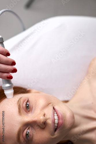 beautiful redhead woman receiving facial microcurrent treatment by beauty therapist, beautician using electrical impulses, equipment. cosmetology, skin care concept