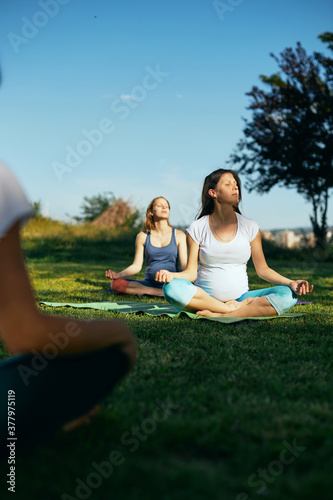 Two calm pregnant women sitting outdoors and meditating while their instructor showing them exercises.