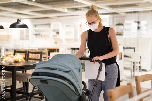 Young mom with newborn in stroller shopping at retail furniture and home accessories store wearing protective medical face mask to prevent spreading of corona virus. New normal during covid epidemic.