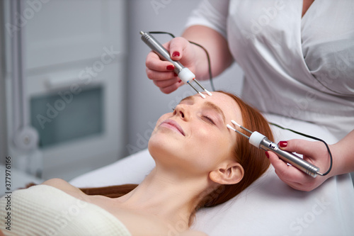 professional beautician with a patient during facial procedure in cosmetological clinic  redhead woman receives an electric facial massage. skin rejuvenation and wrinkle smoothing