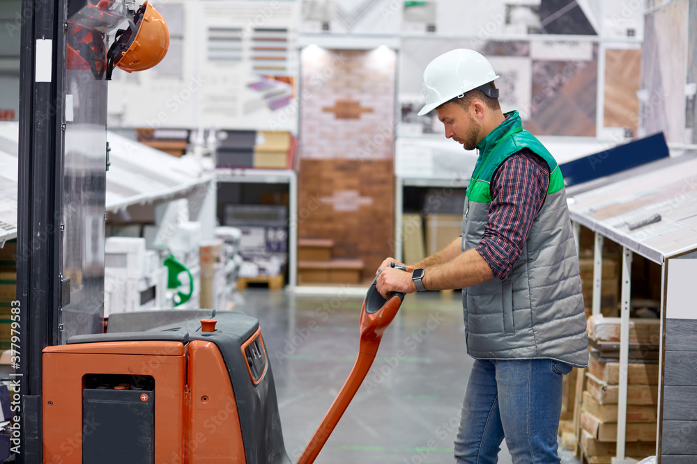 young caucasian warehouse worker with fork pallet truck stacker at modern warehouse, hardworking male at work place, wearing uniform