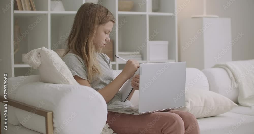 teen girl is viewing online training by laptop from home and learning distantly, writing notes in copybook, relax and domestic atmosphere teen girl is viewing online training by laptop from home and learning distantly, writing notes in copybook, relax and domestic atmosphere
