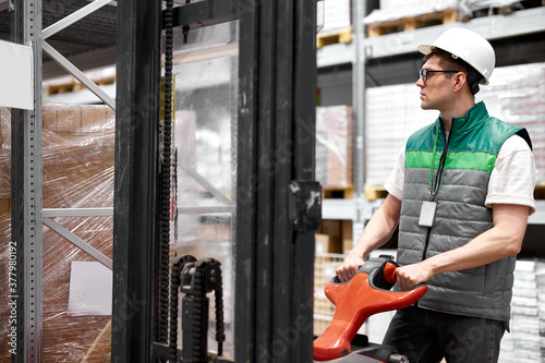young caucasian warehouse worker with fork pallet truck stacker at modern warehouse, hardworking male at work place, wearing uniform