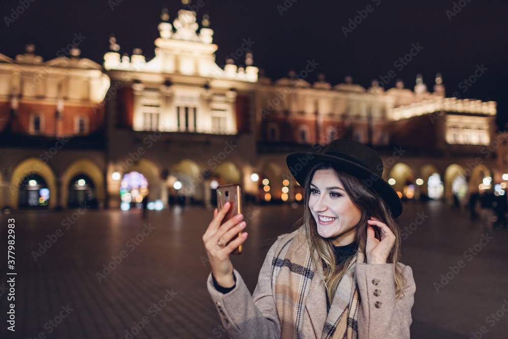 Tourist woman taking selfie on smartphone at night on Market square in Krakow Poland. Travel around Europe