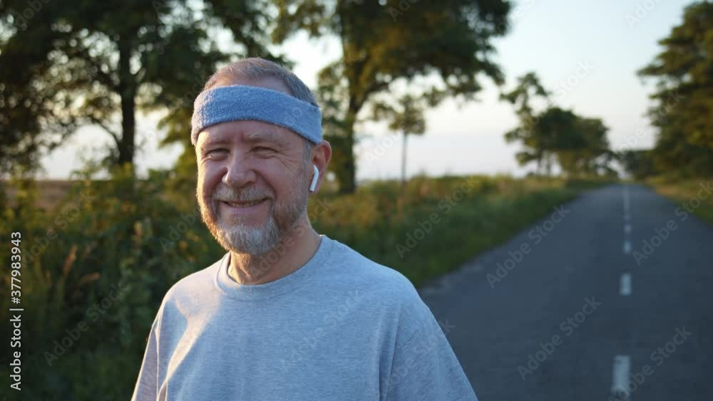 Funny laughing senior man with earphones and head bandage making funny face looking at camera while resting after training outside. Sporty bearded guy enjoying healthy active life. Summer nature. 4K Funny laughing senior man with earphones and head bandage making funny face looking at camera while resting after training outside. Sporty bearded guy enjoying healthy active life. Summer nature. 4K