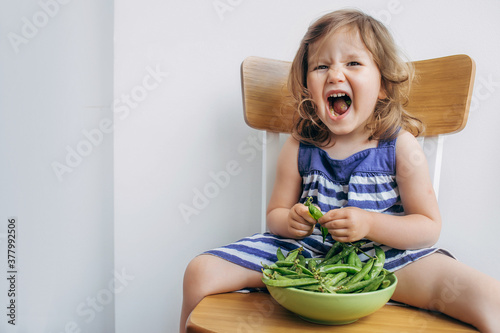 Cheerful little todler girl in a striped dress eating peas and grimacing