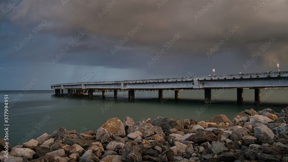 Eine menschenleere Brücke über das Meer in Panama, Steine im Vordergrund und lila Wolken, kurz vor einem Gewitter
