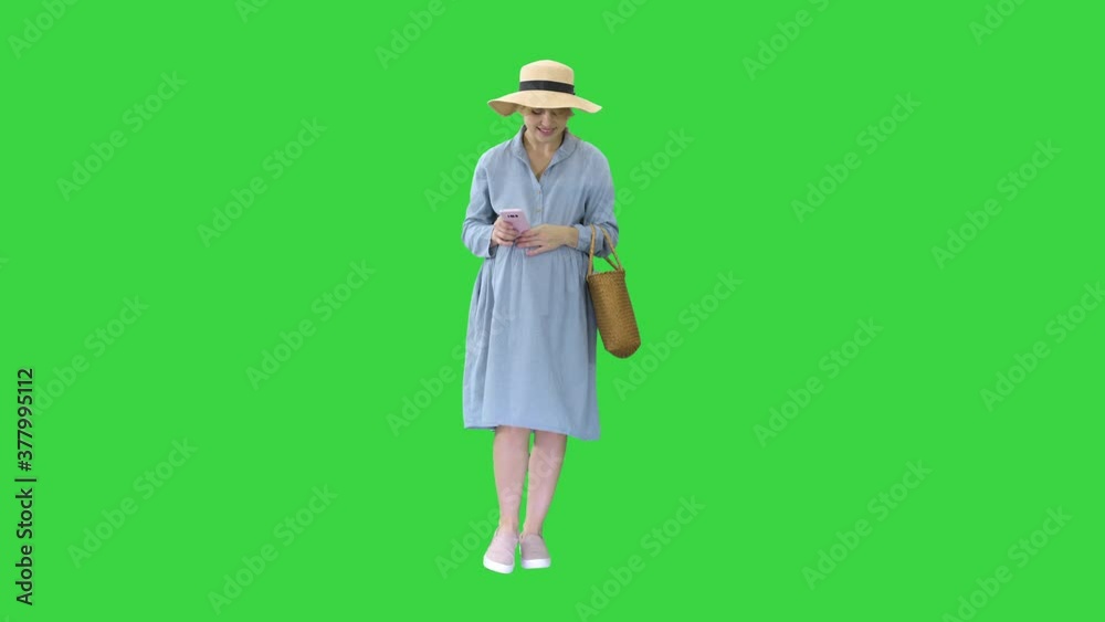 Young pregnant woman in trendy hat walking and using her phone on a Green Screen, Chroma Key.