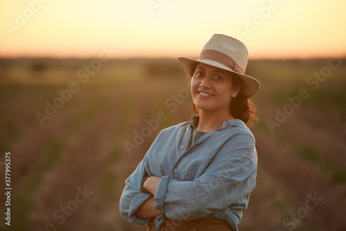 Waist up portrait of young female farmer posing confidently with arms crossed while standing in field at sunset and smiling at camera, copy space