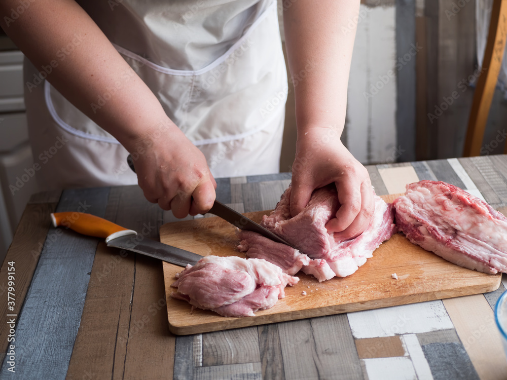 Young extravagant woman cuts large pieces of pork and beef. Fresh meat on cutting board, closeup hands, sharp kitchen knives, white apron