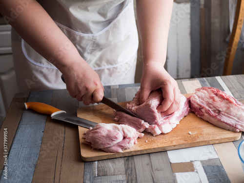 Young extravagant woman cuts large pieces of pork and beef. Fresh meat on cutting board, closeup hands, sharp kitchen knives, white apron