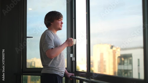 Side view of a happy young manholding cup of coffee on balcony at home in the morning. Concept. Man drinking tea or coffee and standing against an opened window. photo