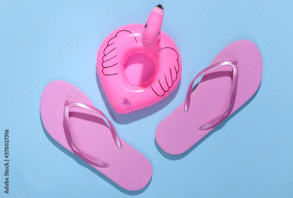 Inflatable pink flamingo with flip flops on blue sunny background. Summer, beach vacation concept. Minimalism. Top view. Inflatable pink flamingo with flip flops on blue sunny background. Summer, beach vacation concept. Minimalism. Top view.