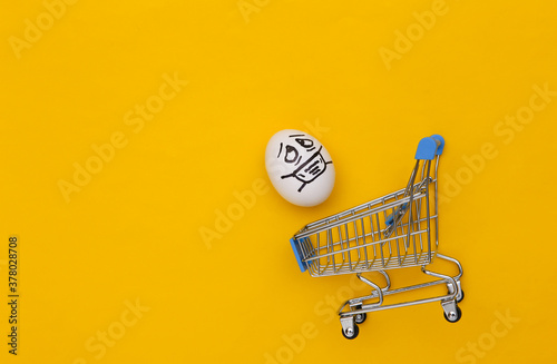 Supermarket trolley with egg face in a medical mask on yellow background. Covid-19 pandemic. Top view photo