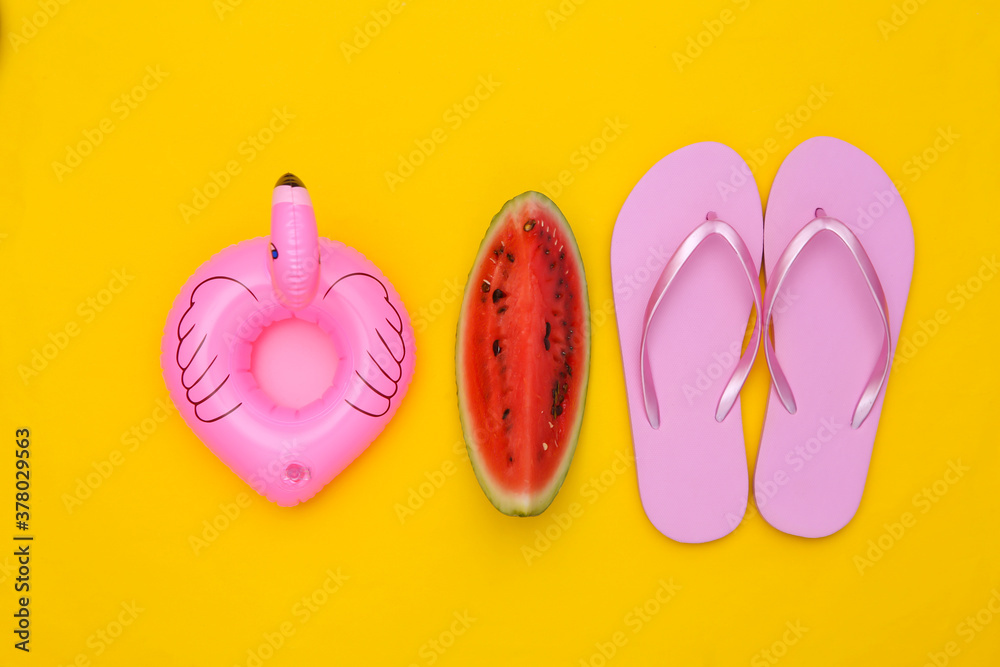 Slice of ripe watermelon and pink flamingo lifebuoy, flip flops on yellow background. Summer fun, beach rest. Top view. Flat lay
